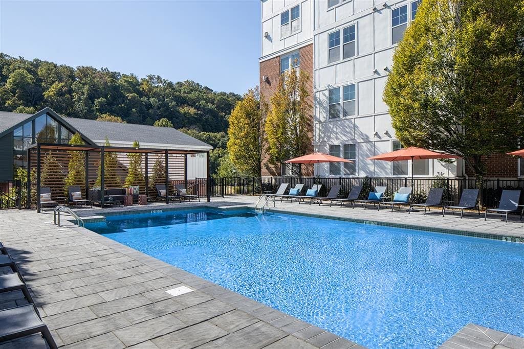 A clear blue swimming pool surrounded by lounge chairs and trees at an apartment complex.