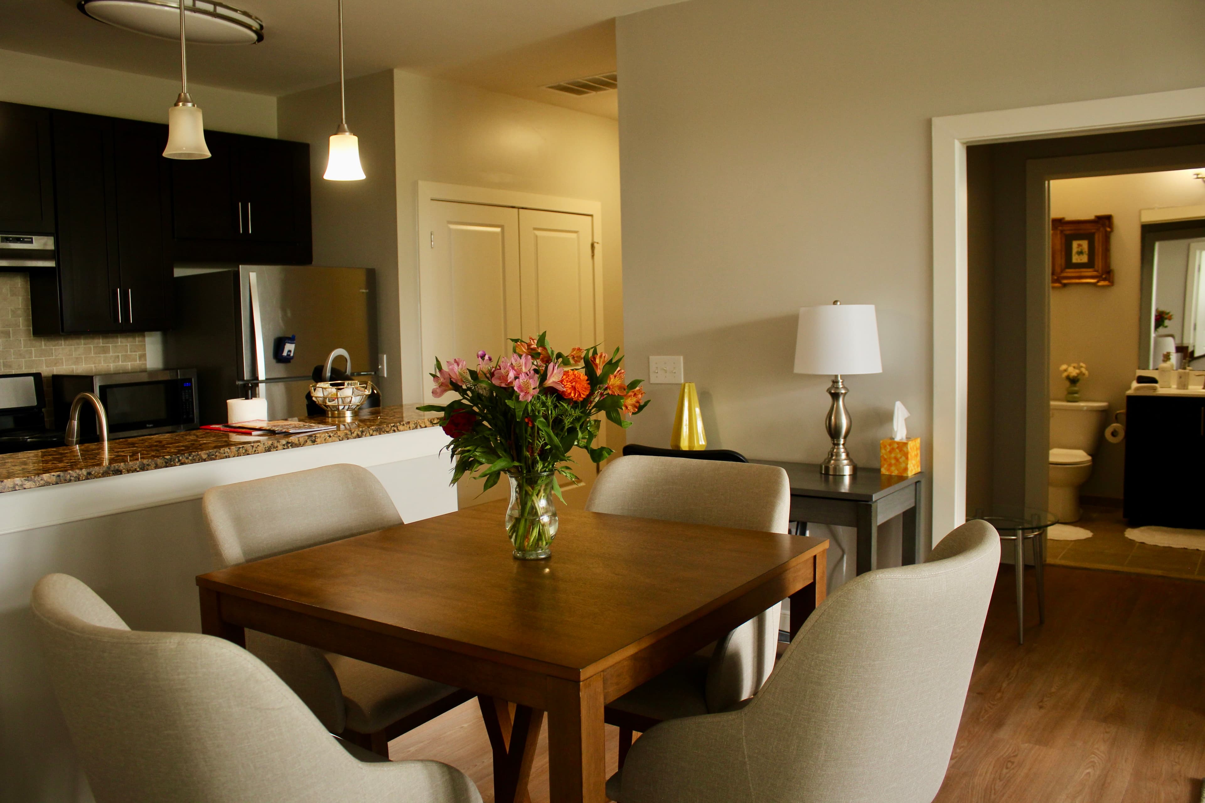 A cozy dining area featuring a wooden table, upholstered chairs, and a bouquet of flowers, with an open kitchen and bathroom visible in the background.