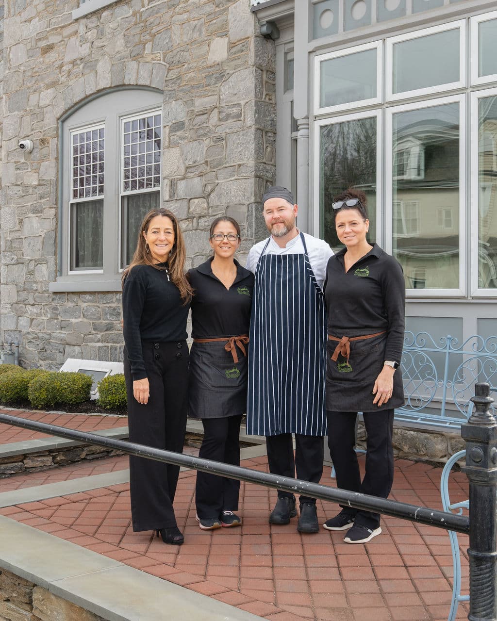 A group of four restaurant staff members, including a chef, stand together outside a stone building.