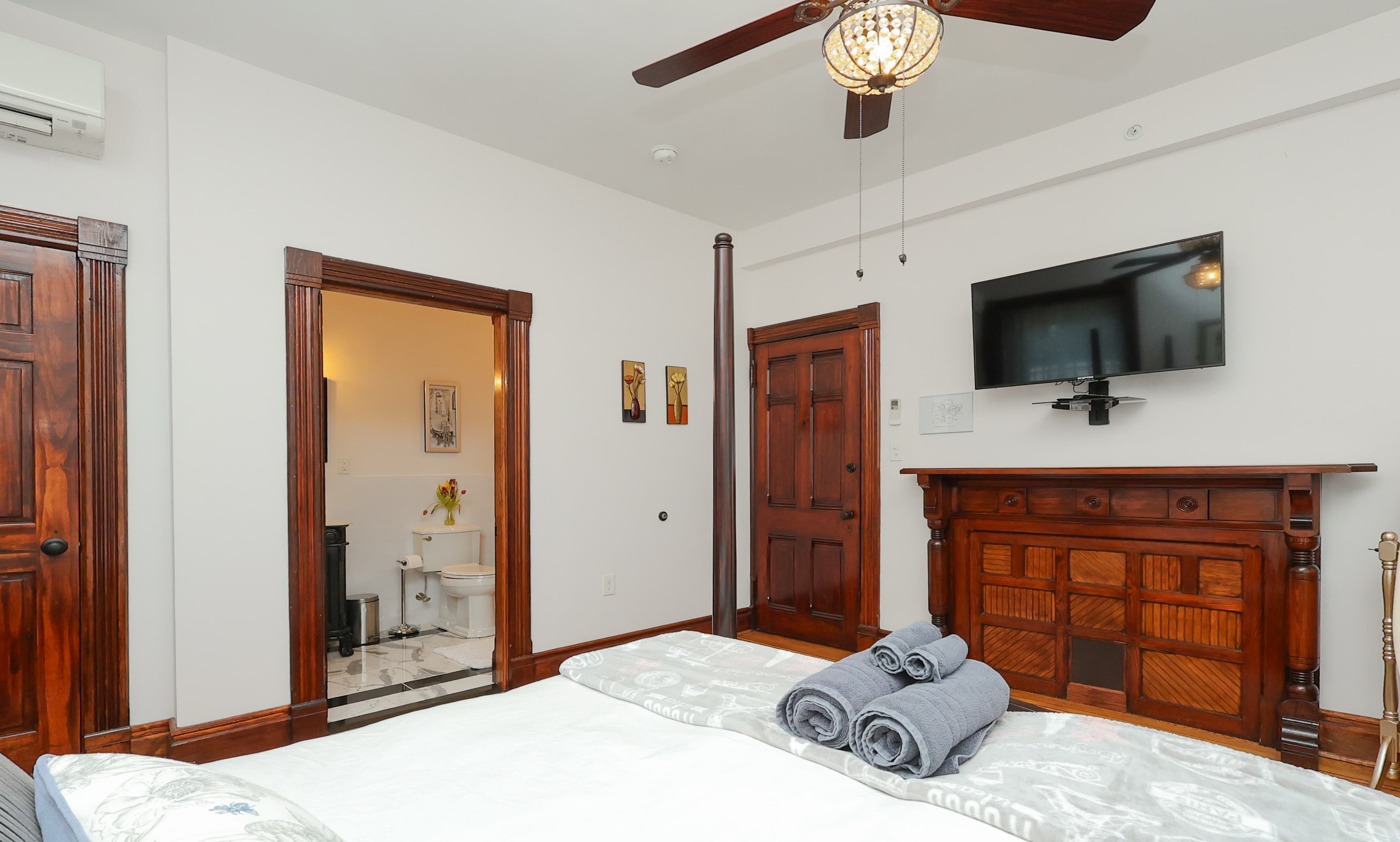 A cozy bedroom featuring a bed with rolled towels, a wall-mounted TV, and a doorway leading to a bathroom.