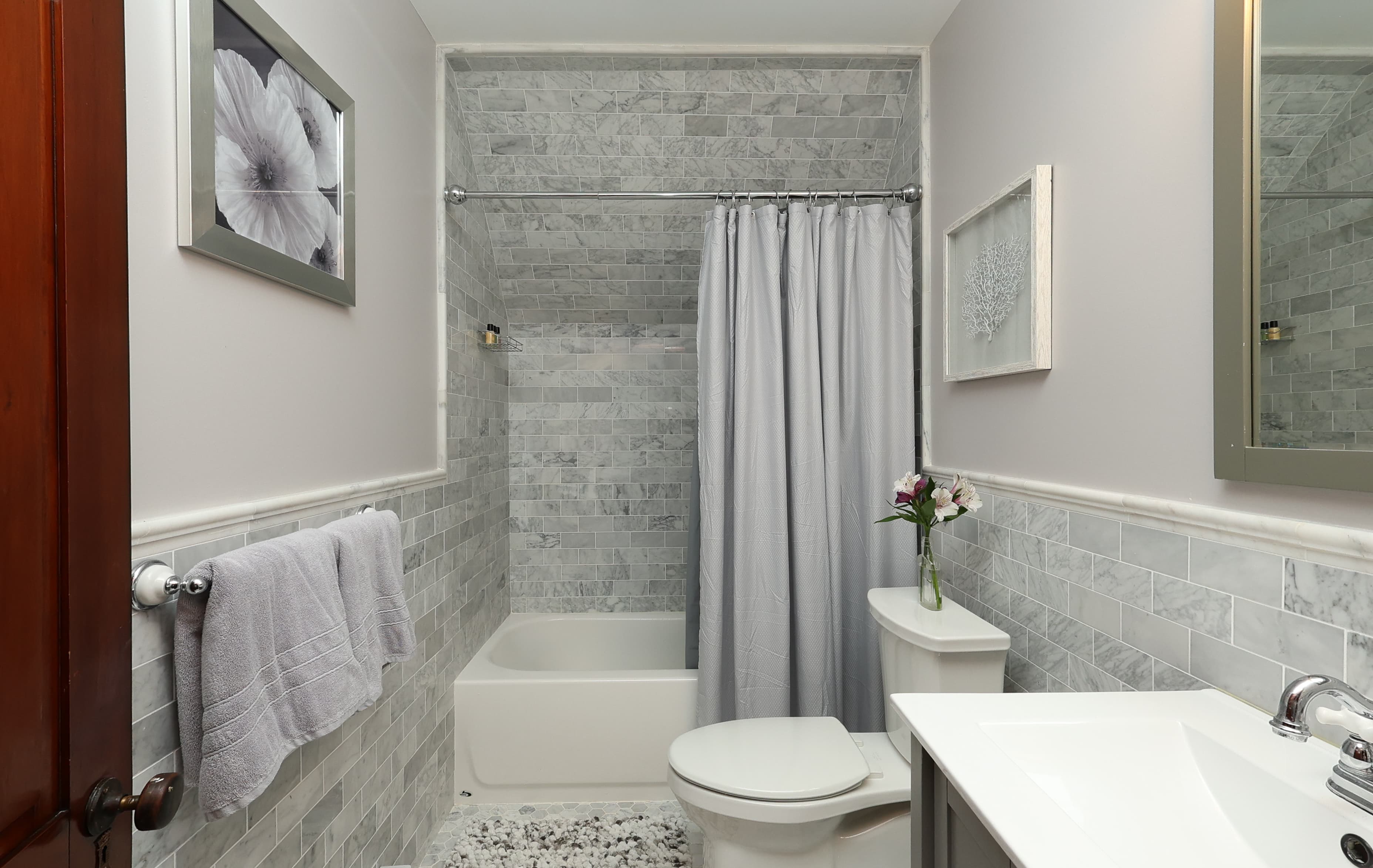 A modern bathroom featuring gray tiled walls, a bathtub with a shower curtain, a white toilet, and a sink with flowers.