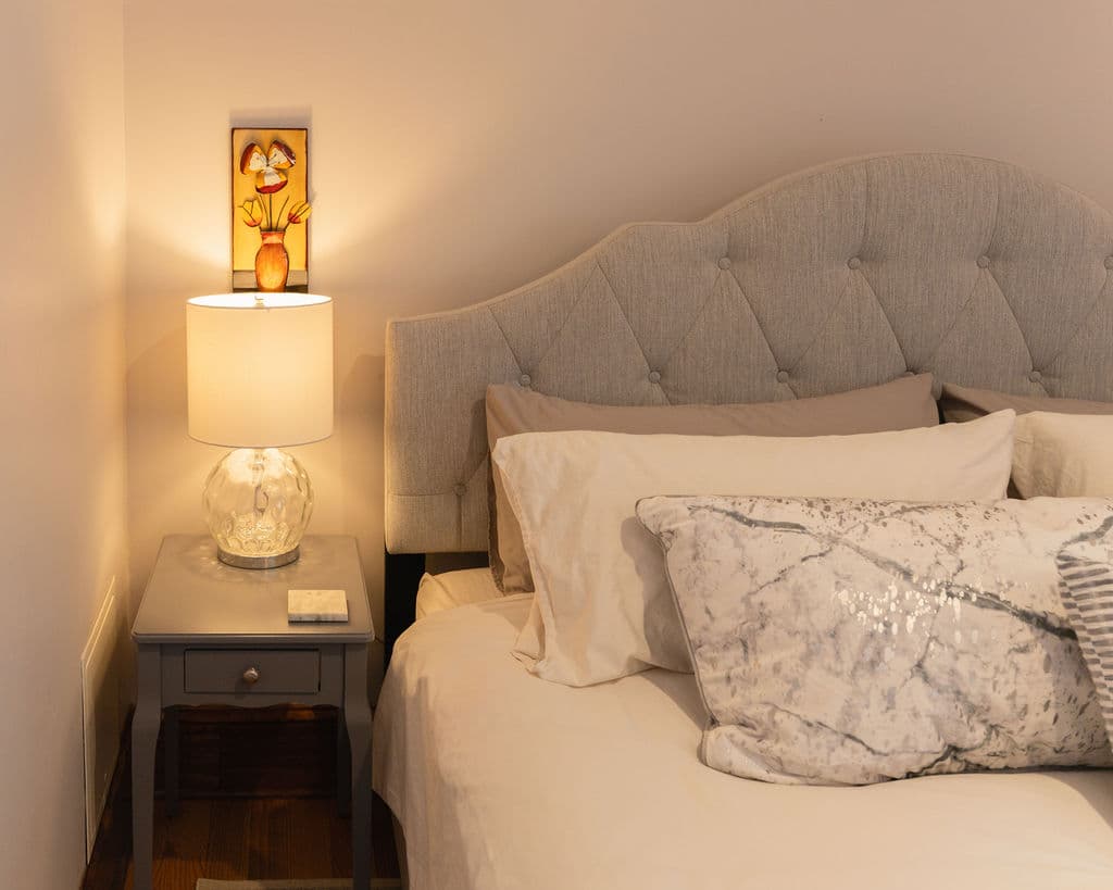 A cozy bedroom corner featuring a tufted headboard, fluffy pillows, a bedside table with a lamp, and a decorative wall sconce.