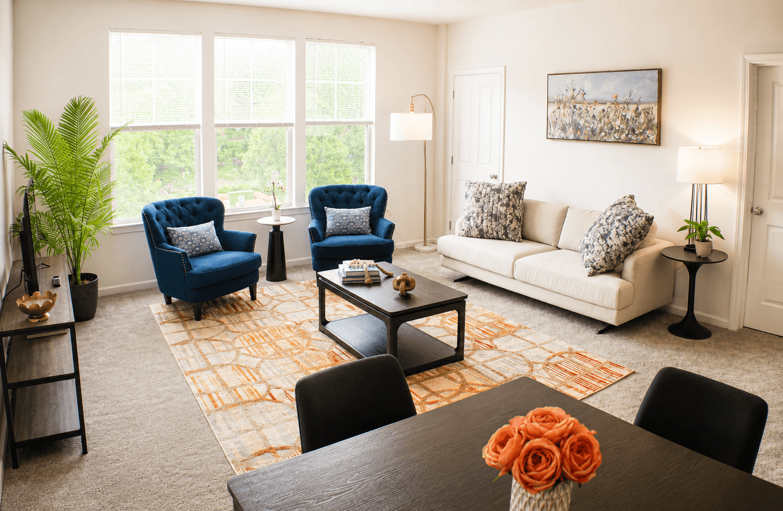 A bright living room featuring a beige sofa, two blue armchairs, a coffee table, and a dining table with a vase of roses.