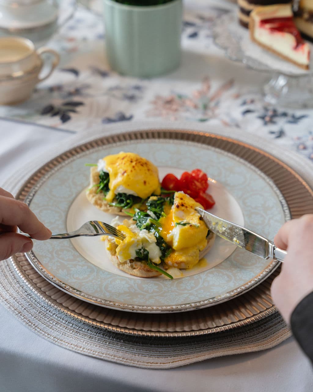 A hand cuts into a plate of eggs Benedict with spinach and cherry tomatoes on a floral tablecloth.