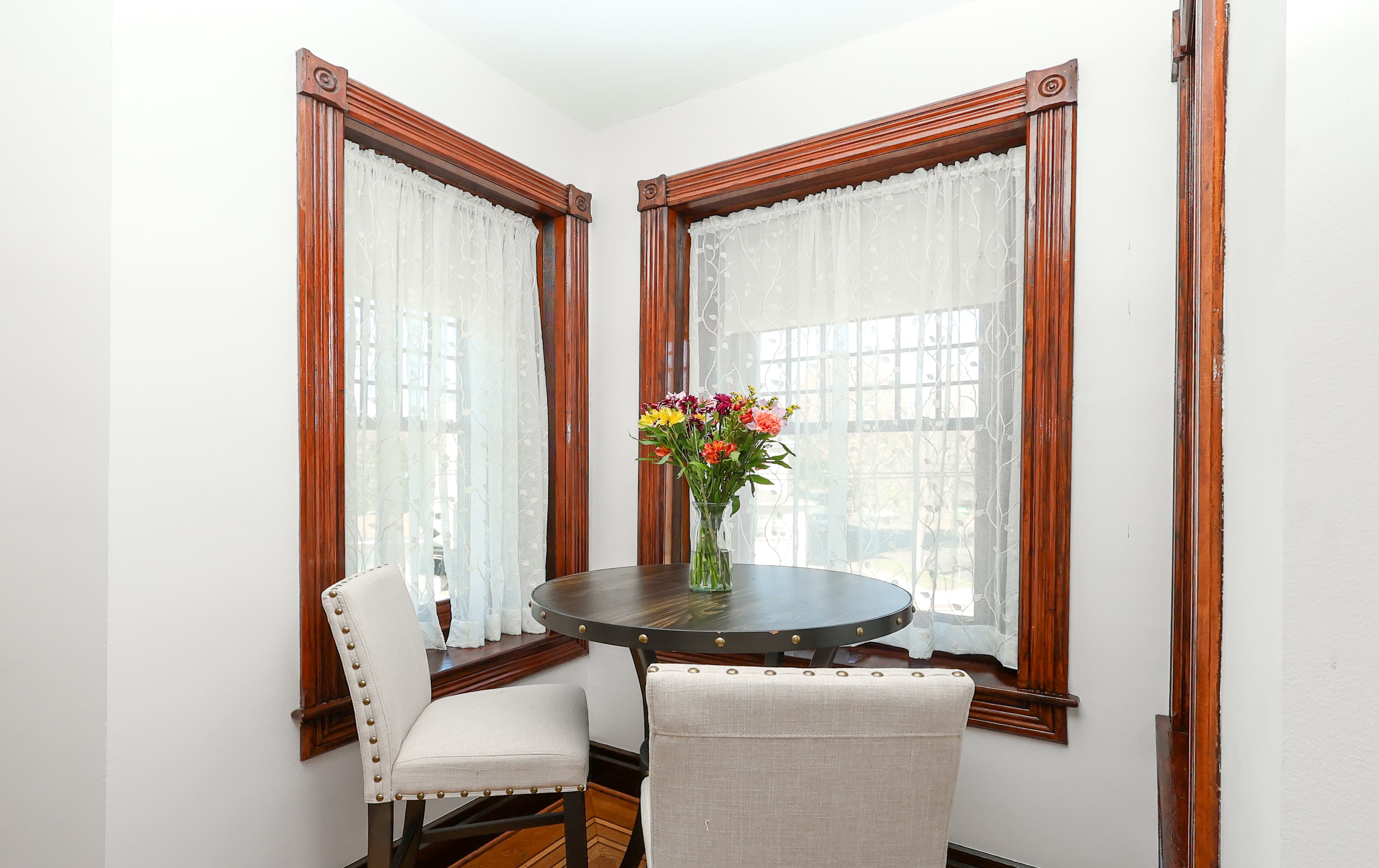 A cozy corner with a round table, two chairs, and a vase of flowers by windowed walls.