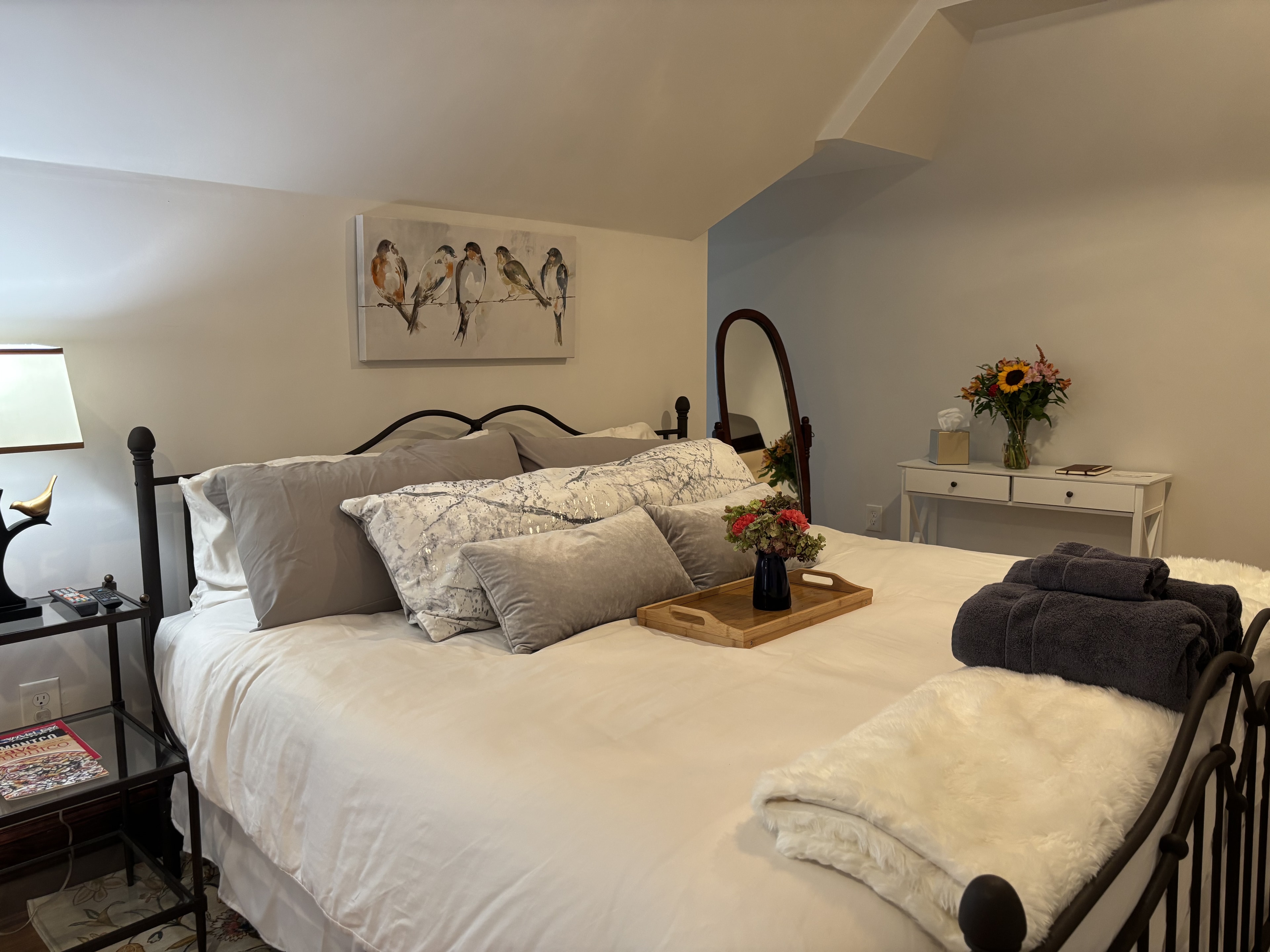 A cozy bedroom featuring a neatly made bed with decorative pillows, a bedside lamp, a mirror, and a small table with flowers.