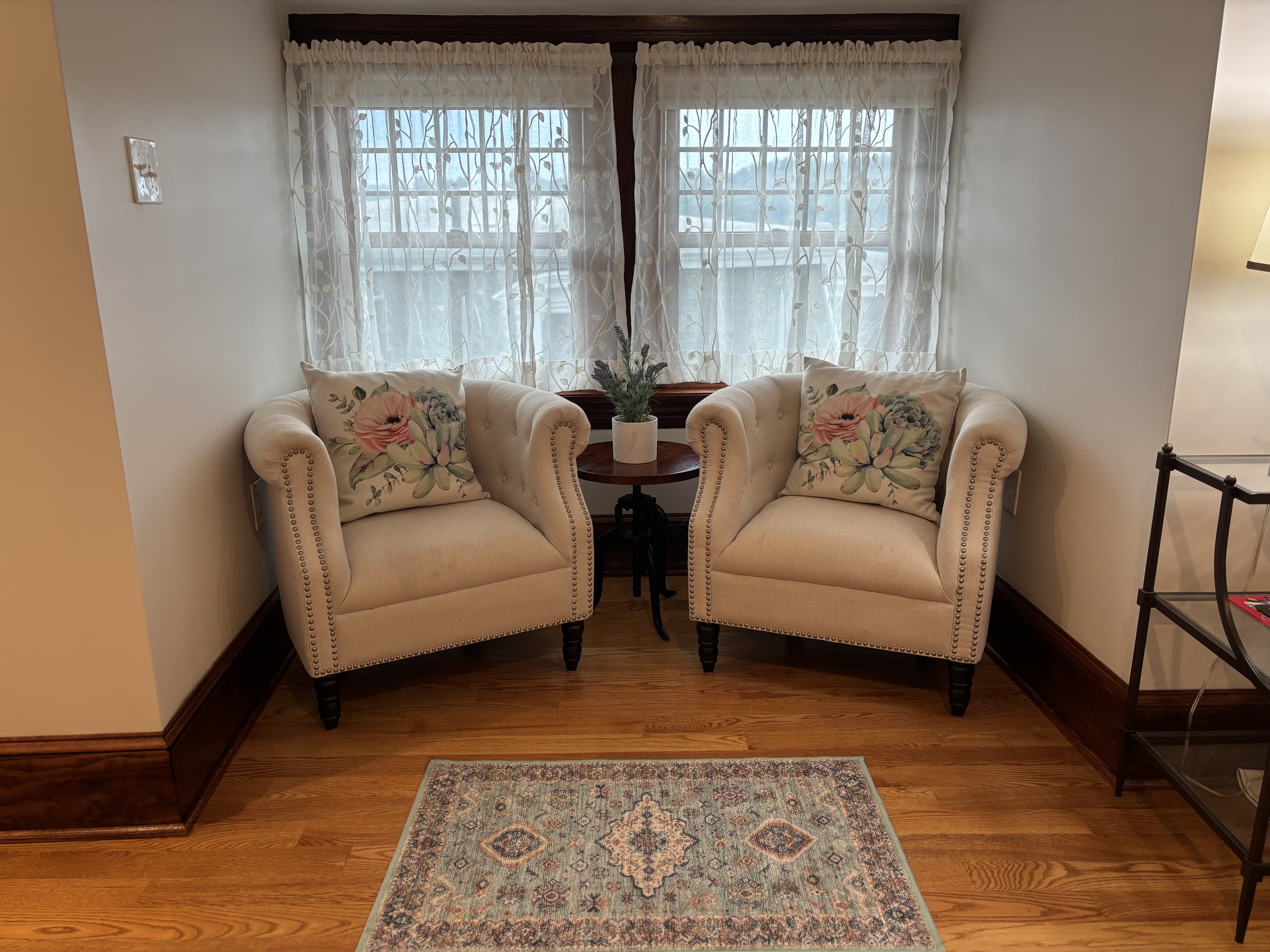 A cozy reading nook with two upholstered armchairs and a small table between them, framed by large windows and lace curtains.