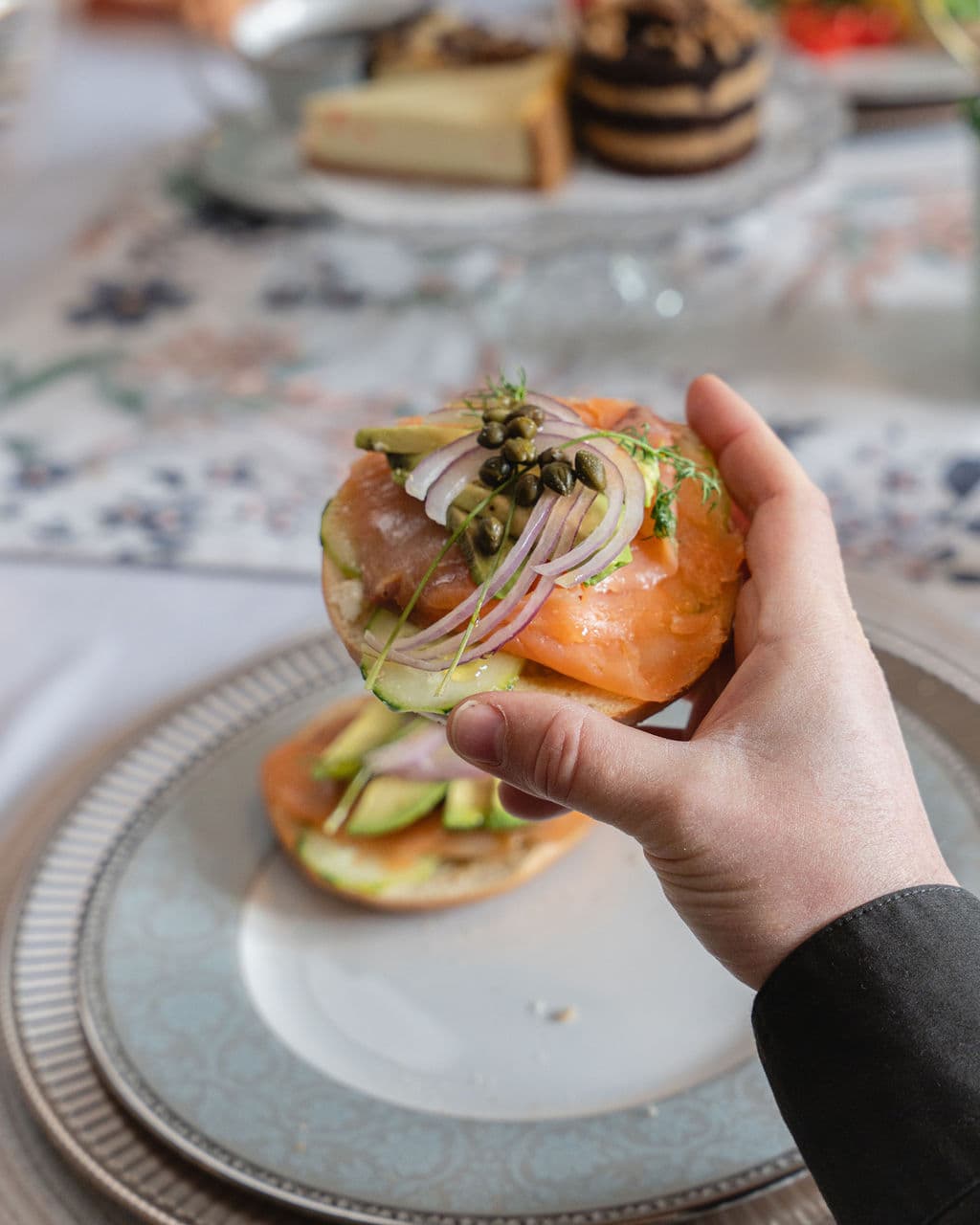 A hand holds a bagel topped with smoked salmon, onions, capers, and herbs, with additional bagel slices on a plate in the background.