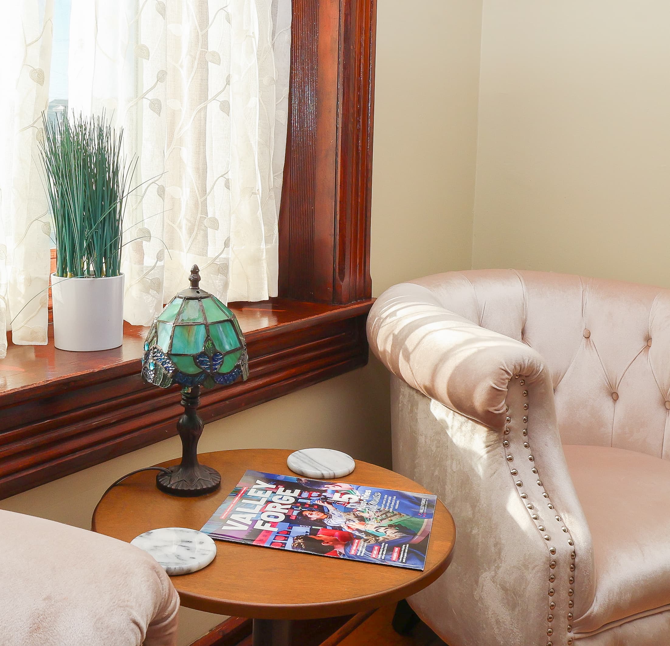 A cozy corner featuring a tufted armchair, a small table with a magazine and coasters, a stained glass lamp, and potted grass by the window.