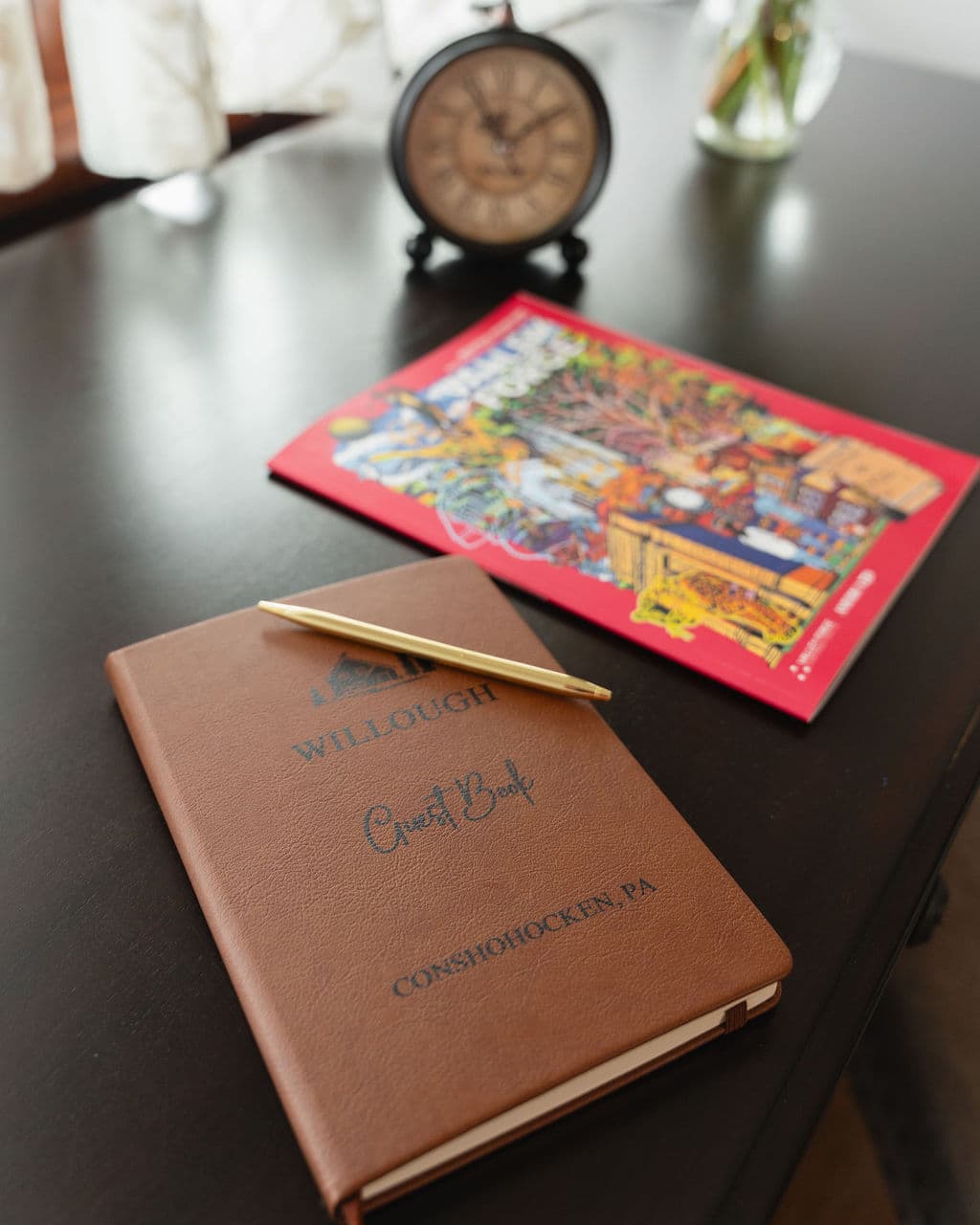 A brown guestbook with a gold pen sits on a table next to a colorful magazine and a clock.