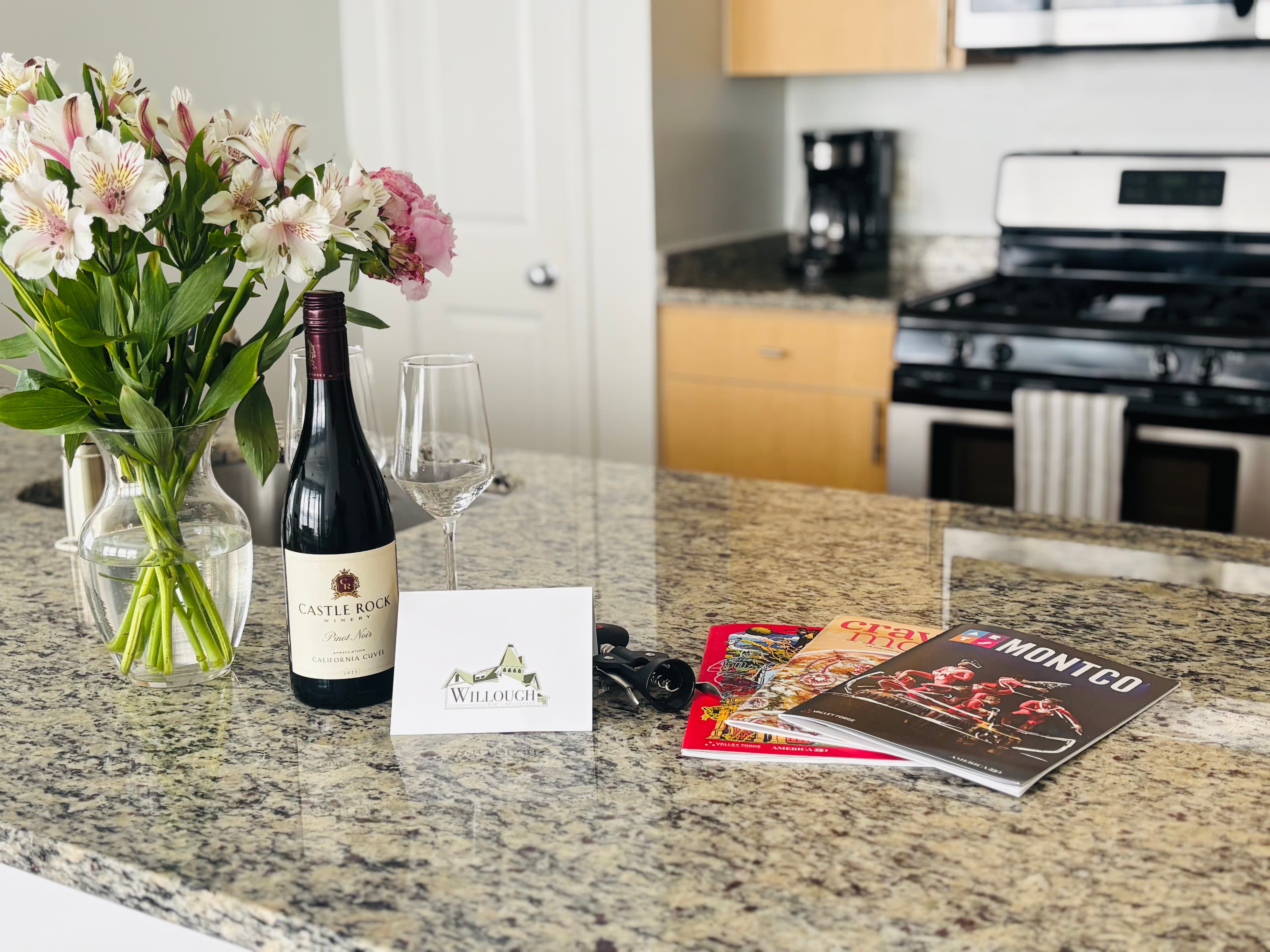 A kitchen countertop with a vase of flowers, a bottle of wine, a glass, a greeting card, keys, and several magazines.