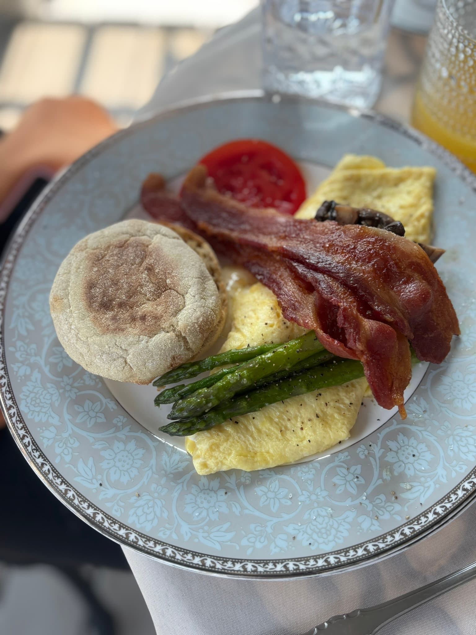 A plate of breakfast featuring bacon, an omelet, asparagus, a tomato slice, and an English muffin.