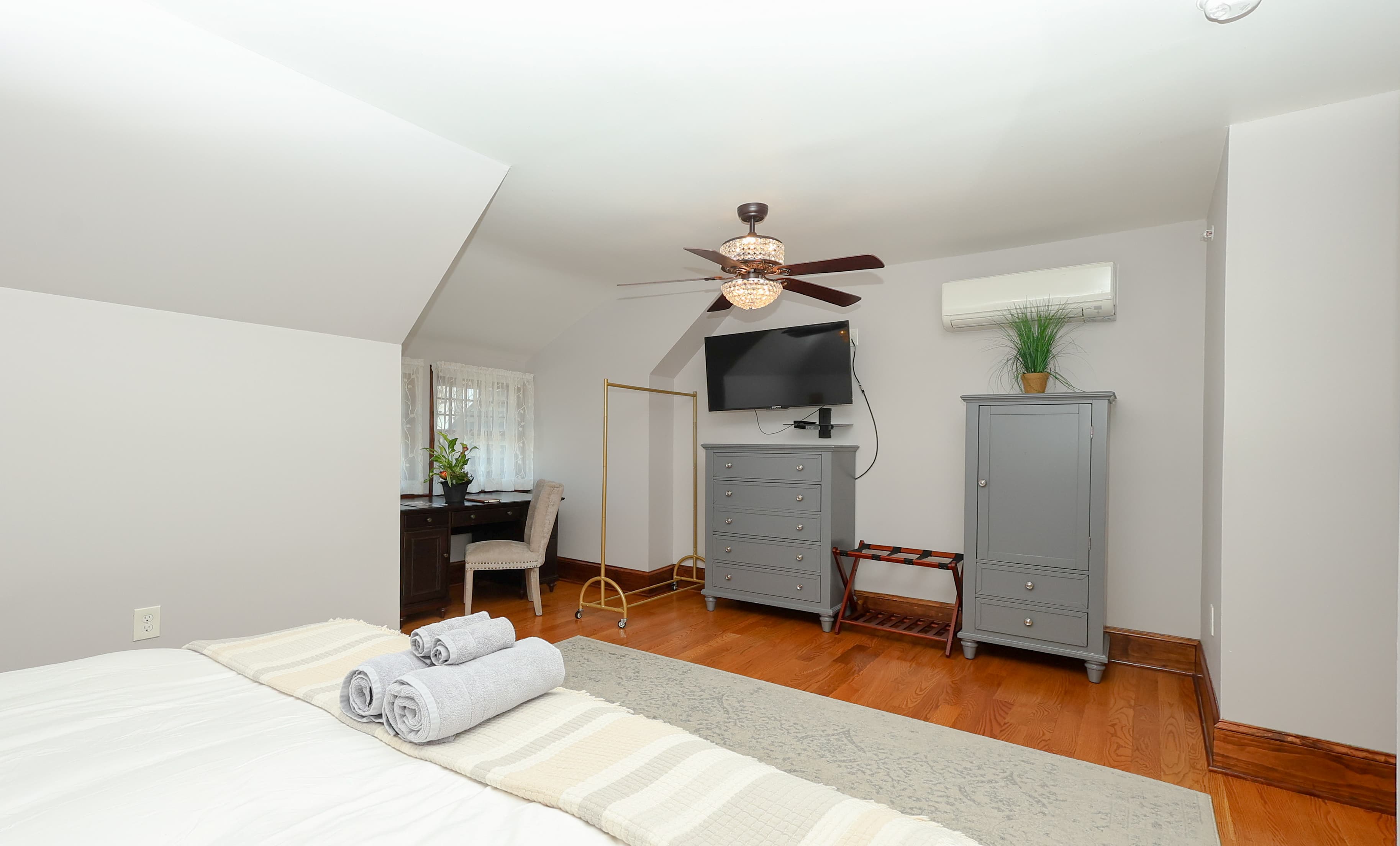 A cozy bedroom featuring a bed with towels, a dresser, a TV, a workspace, and a ceiling fan.