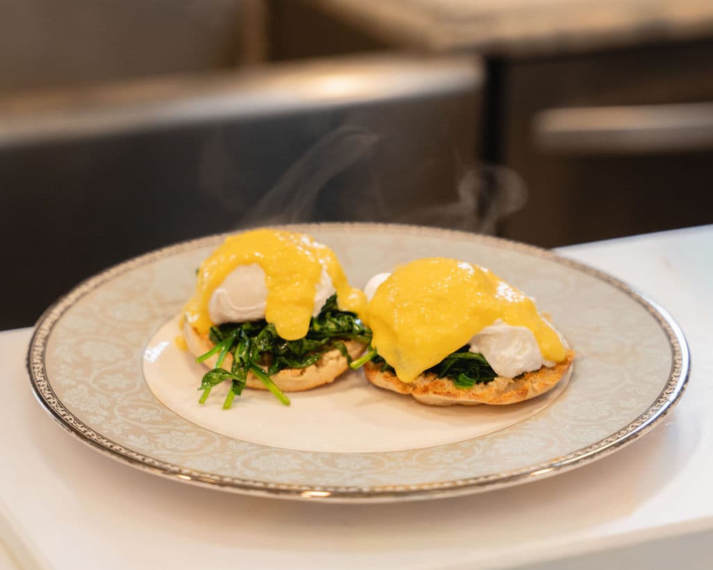 Two poached eggs on toasted English muffins, topped with hollandaise sauce and served with sautéed spinach, emitting steam.