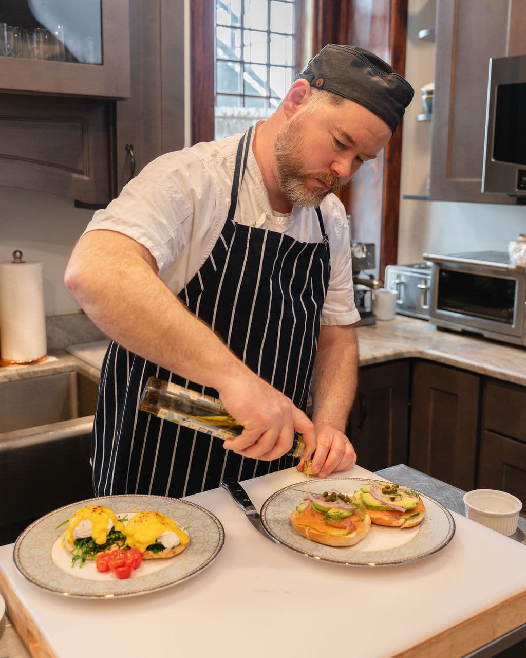 A chef in a striped apron drizzles olive oil over two plated dishes in a kitchen.