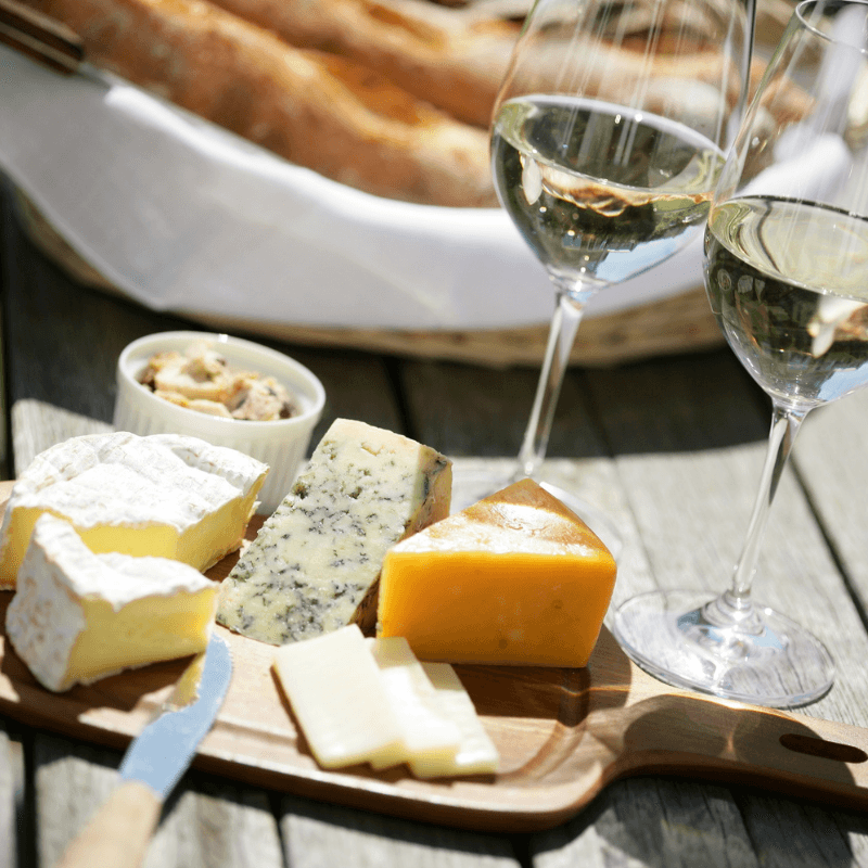 A wooden board displays an assortment of cheeses alongside two glasses of white wine, with a basket of bread in the background.