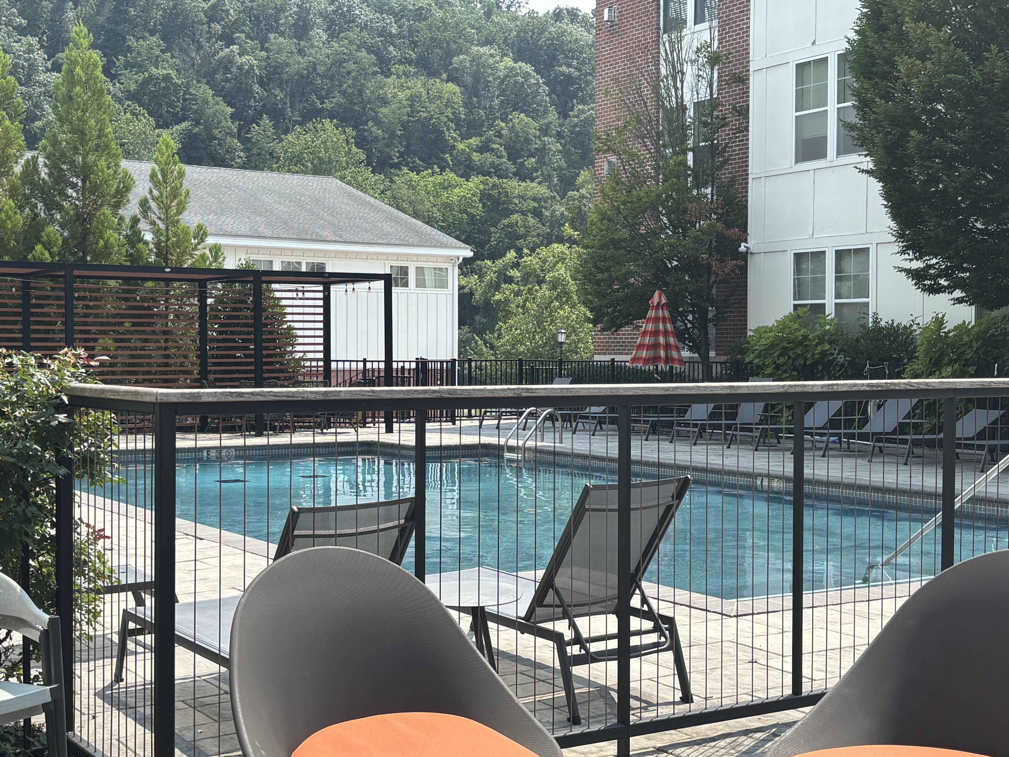 A swimming pool surrounded by lounge chairs and greenery, with an apartment building in the background.