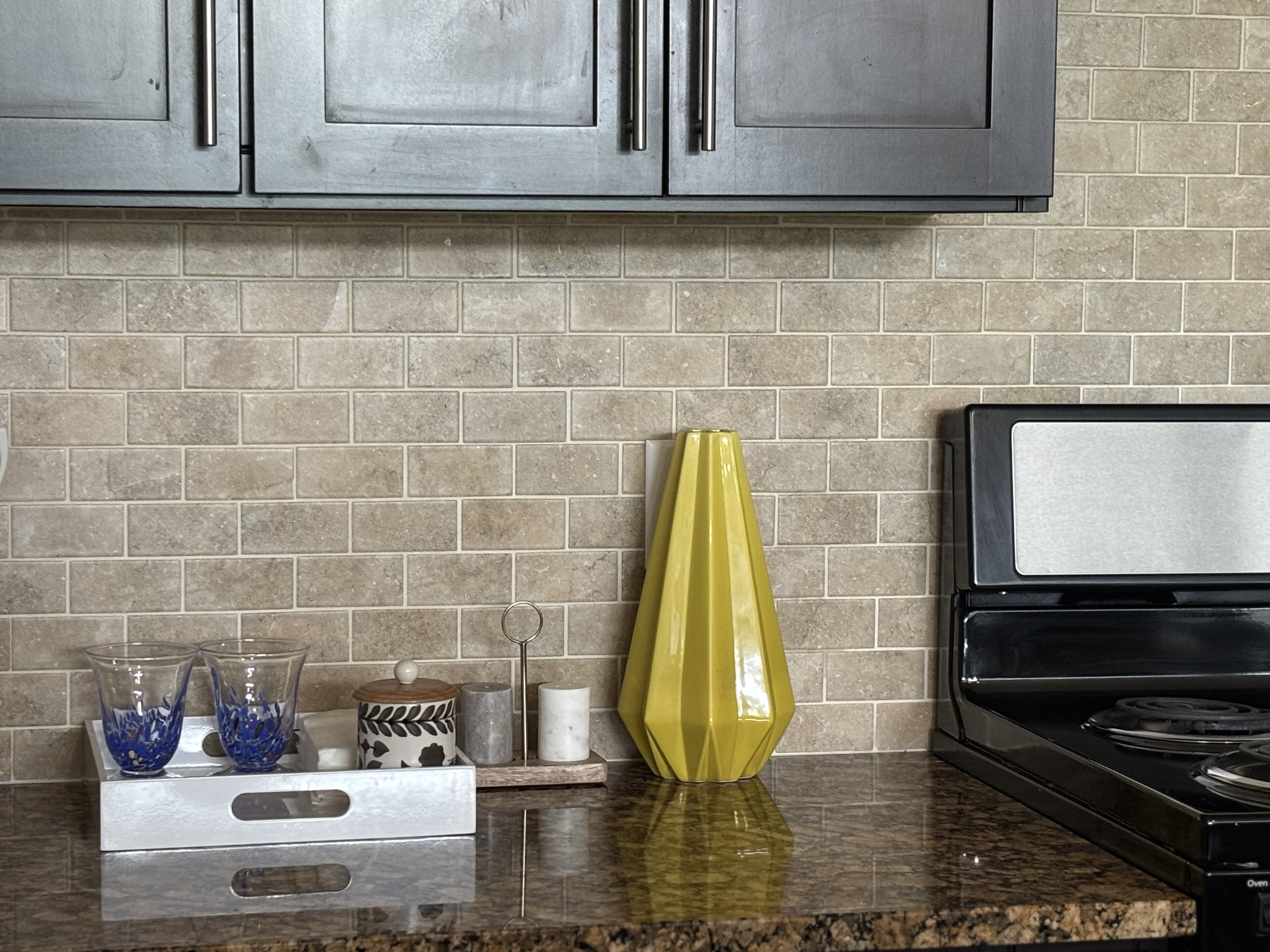 A modern kitchen countertop featuring a yellow vase, decorative items, and glassware against a tiled backsplash.