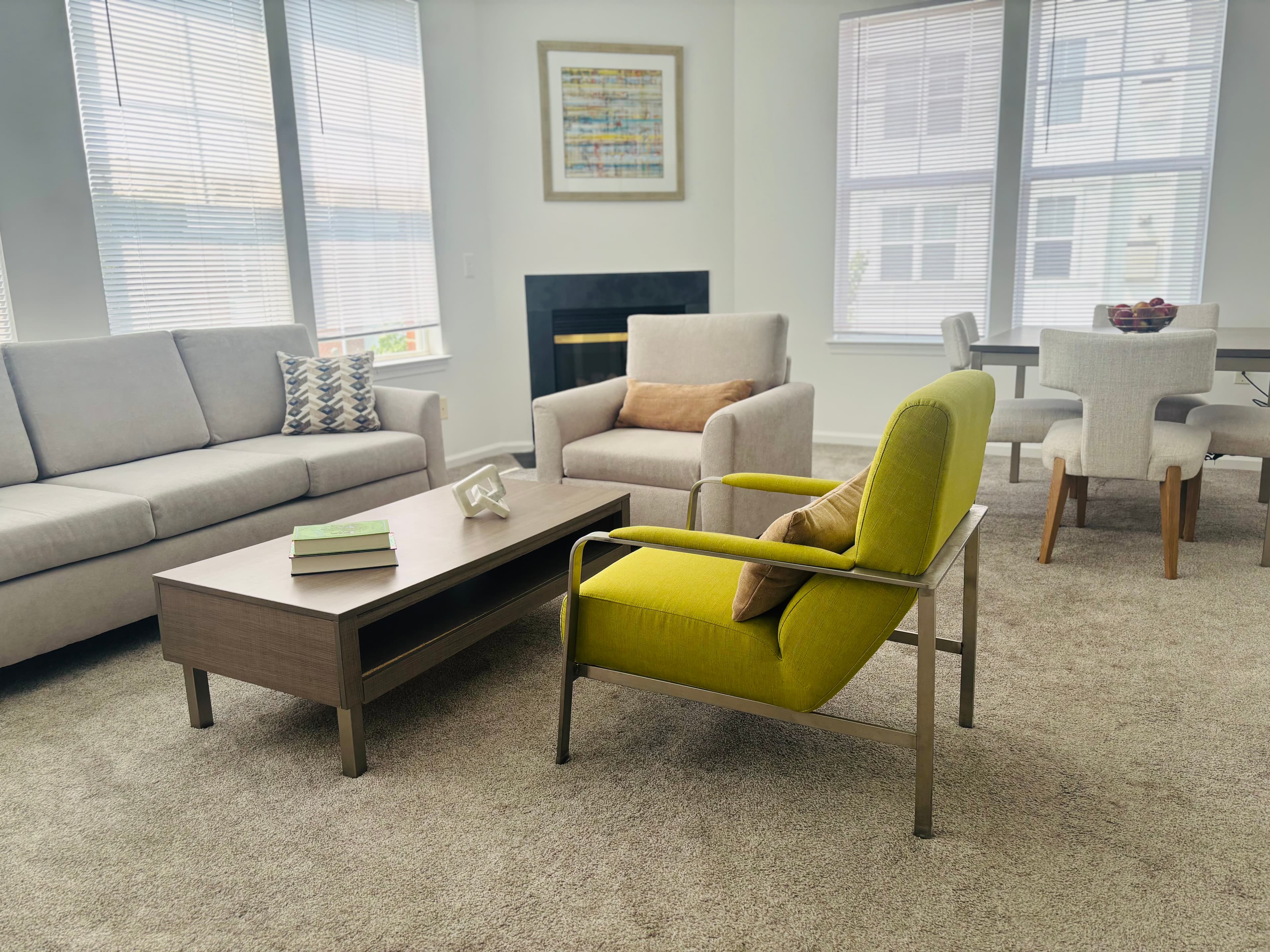 A cozy living room with a gray sofa, a bright green chair, a wooden coffee table with books, and a dining area in the background.