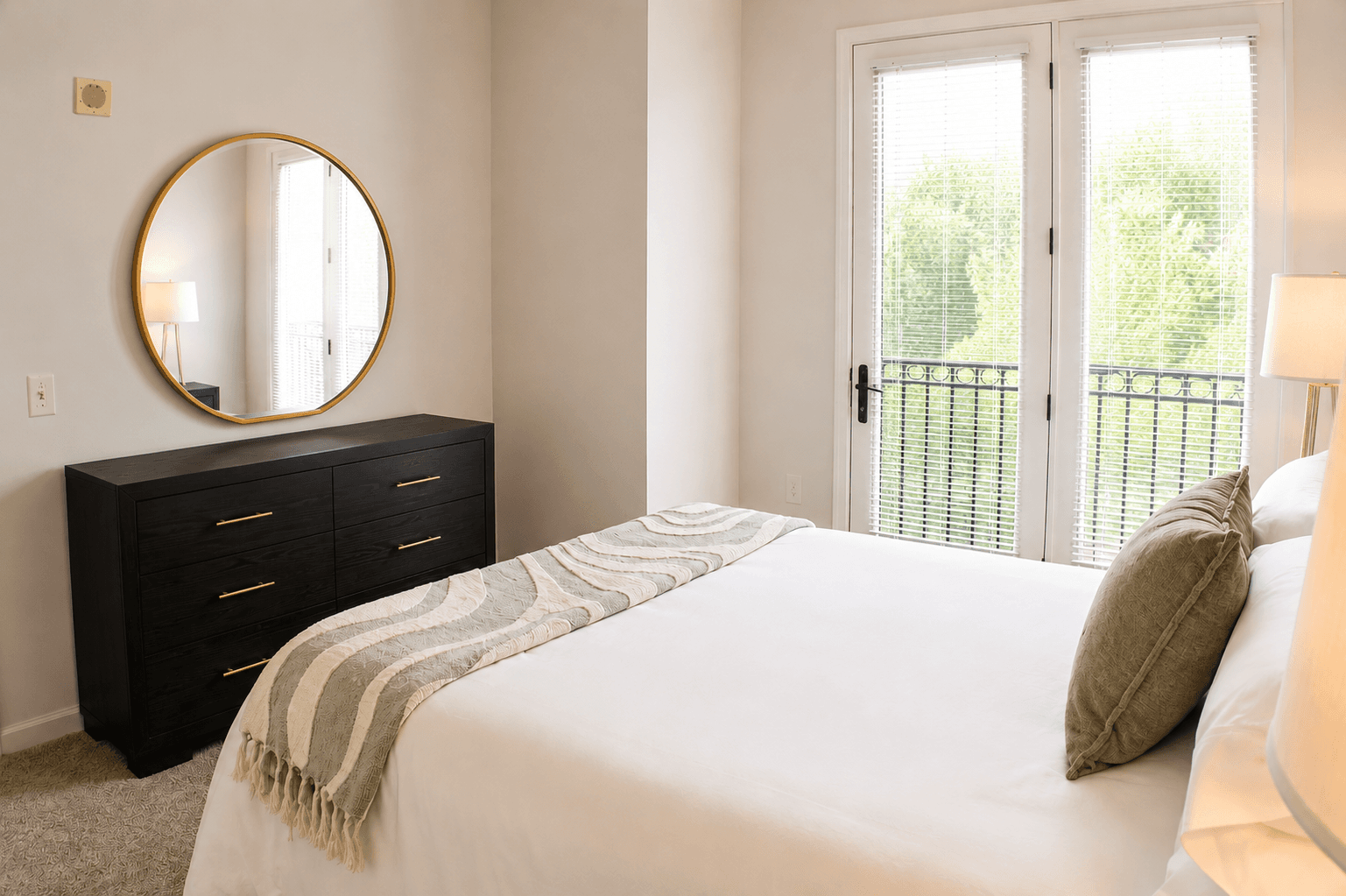 Bright bedroom featuring a white bed, green accent pillow, a round mirror, and large windows with a balcony view.