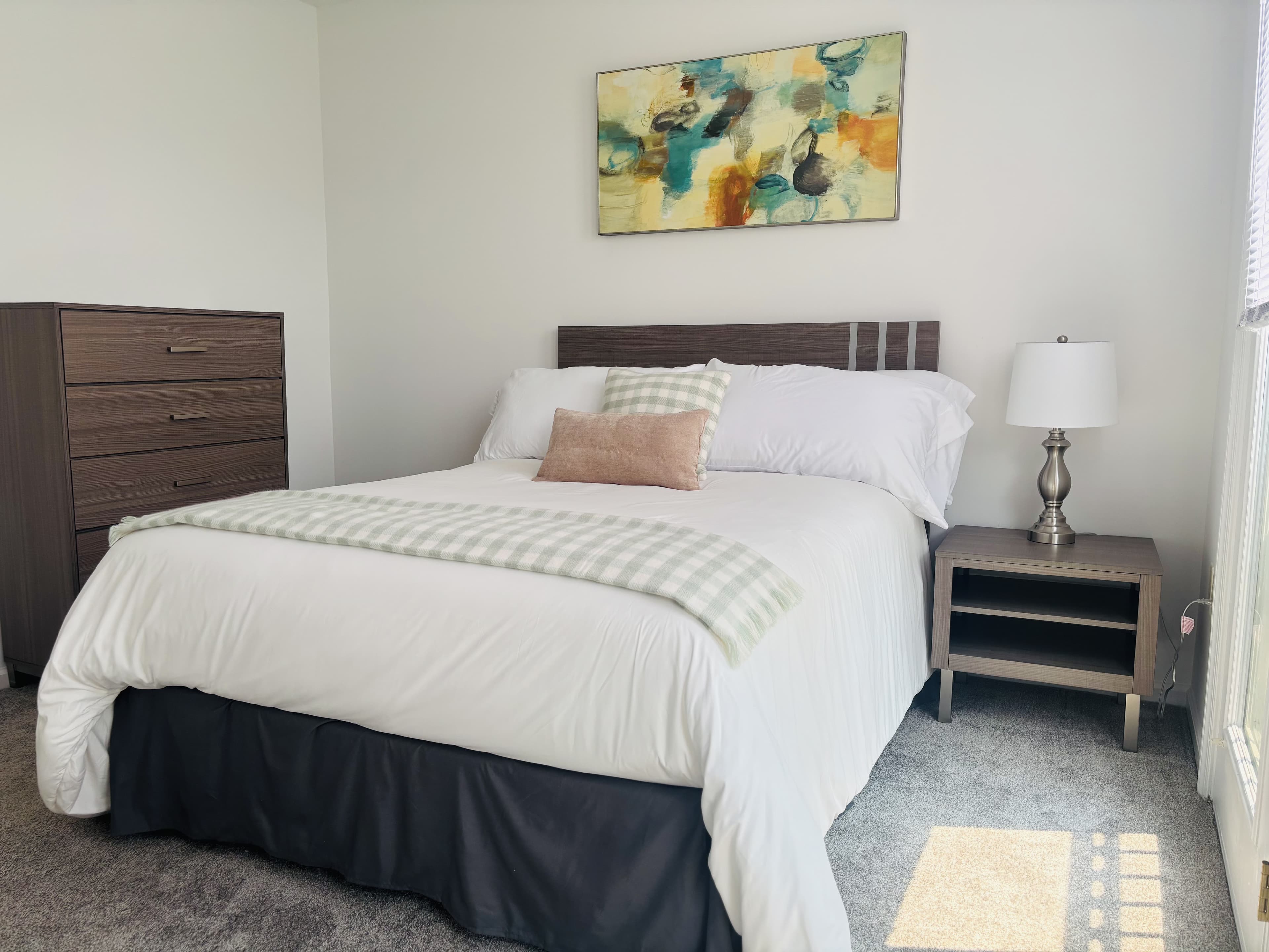 A neatly made bed with a decorative pillow in a bright, modern bedroom featuring a nightstand and a dresser.