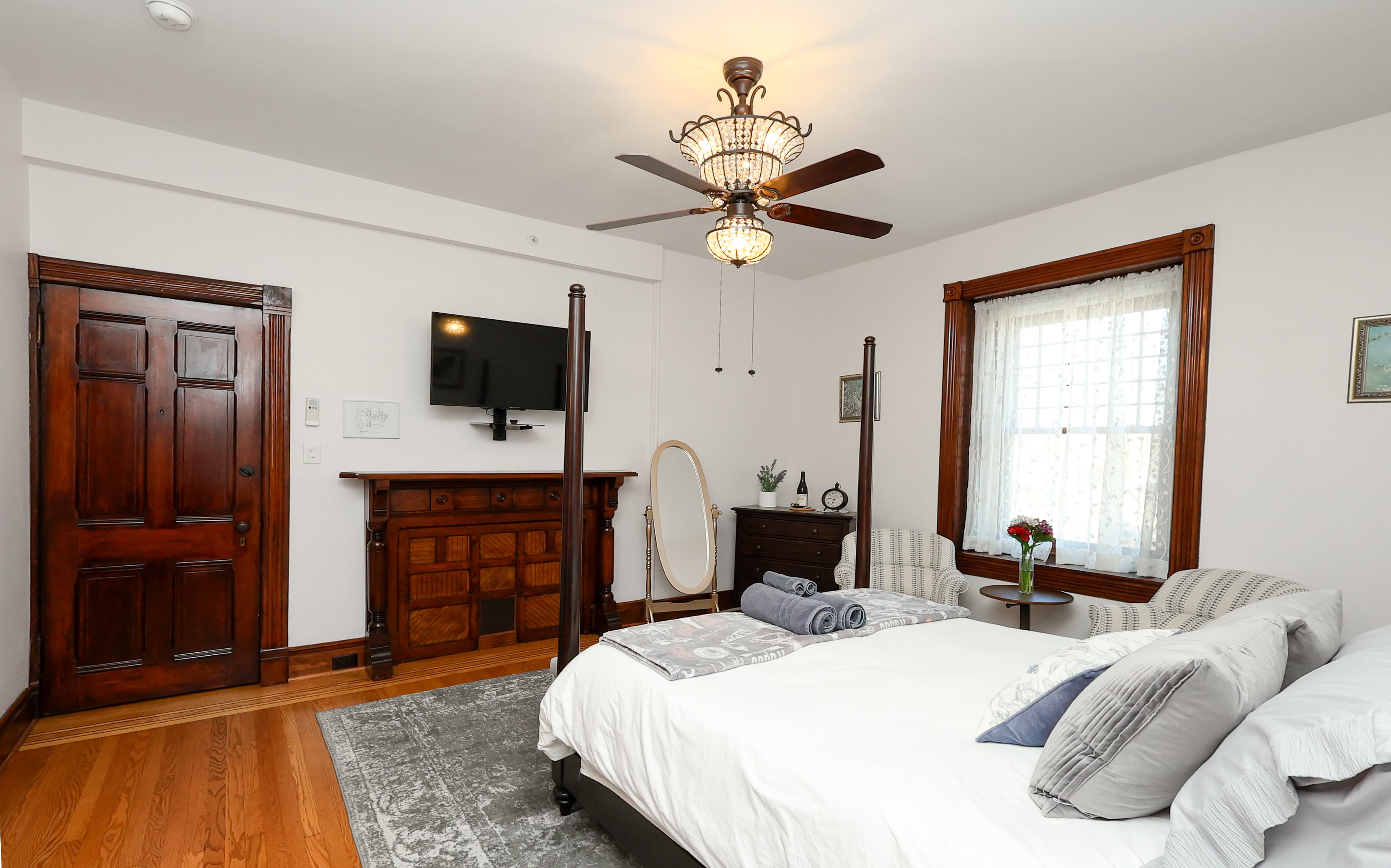 A cozy bedroom featuring a four-poster bed, wooden furnishings, a TV, and natural light from a window.