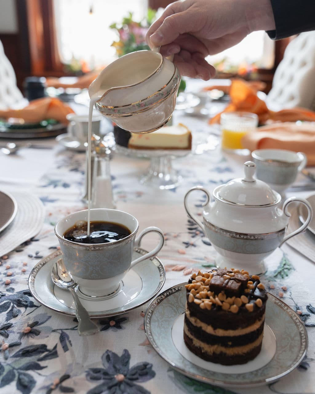 A hand pours cream from a delicate pitcher into a cup of coffee, accompanied by a slice of cake on an elegantly set table.