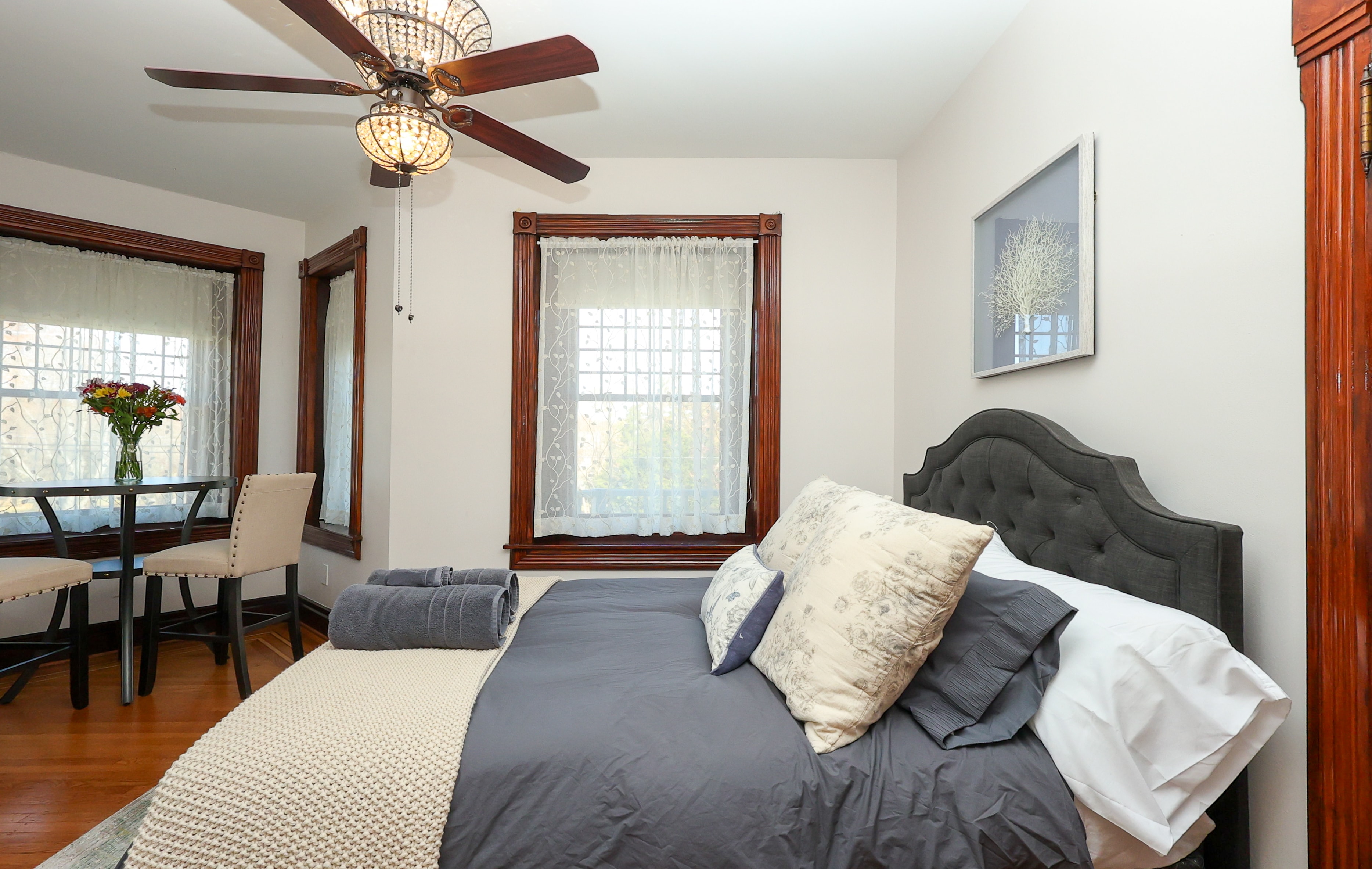 A cozy bedroom with a grey bedspread, decorative pillows, and a small dining table beside two windows.