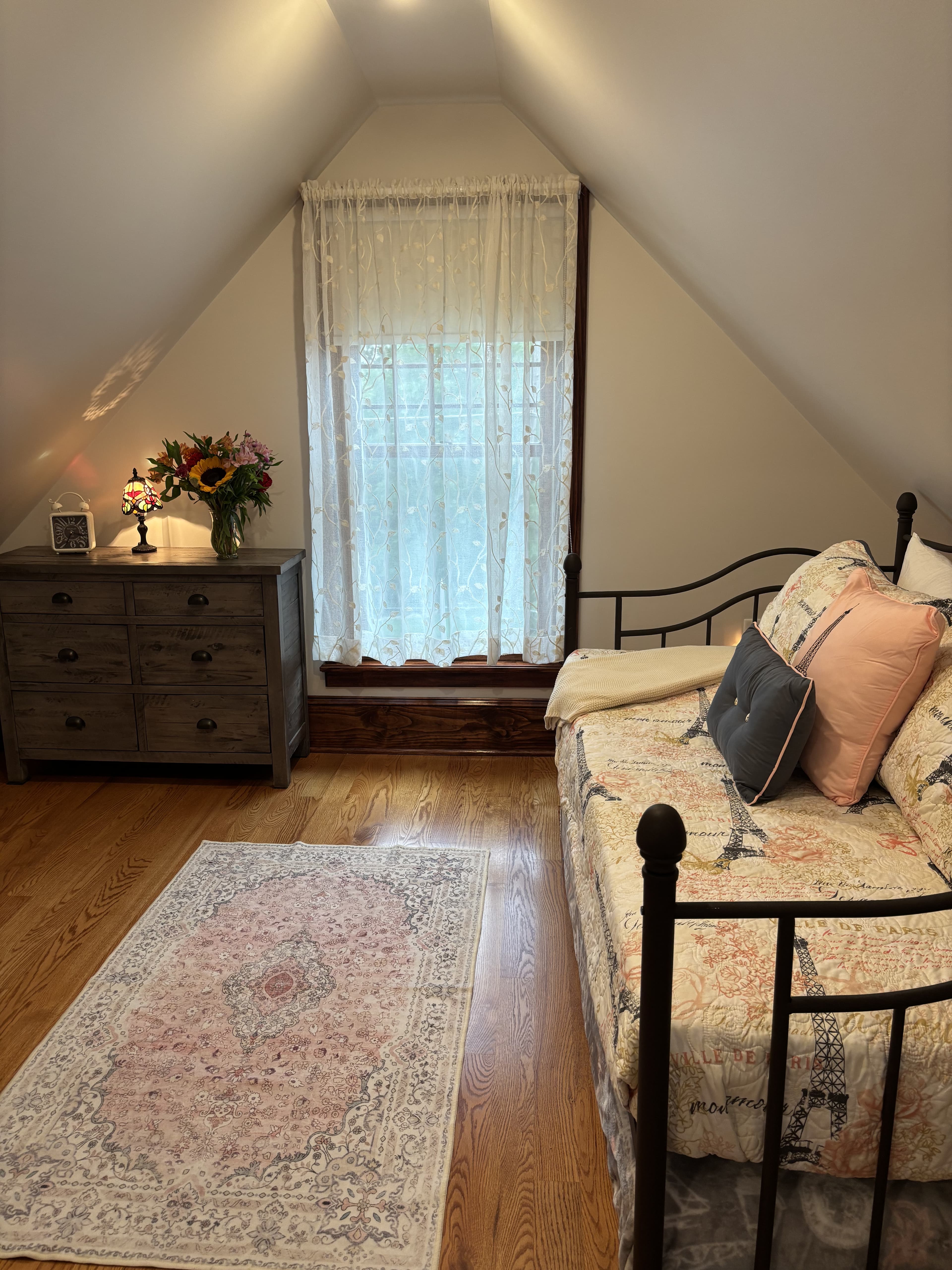A cozy attic bedroom featuring a metal daybed, wooden dresser, floral curtains, and a decorative rug.