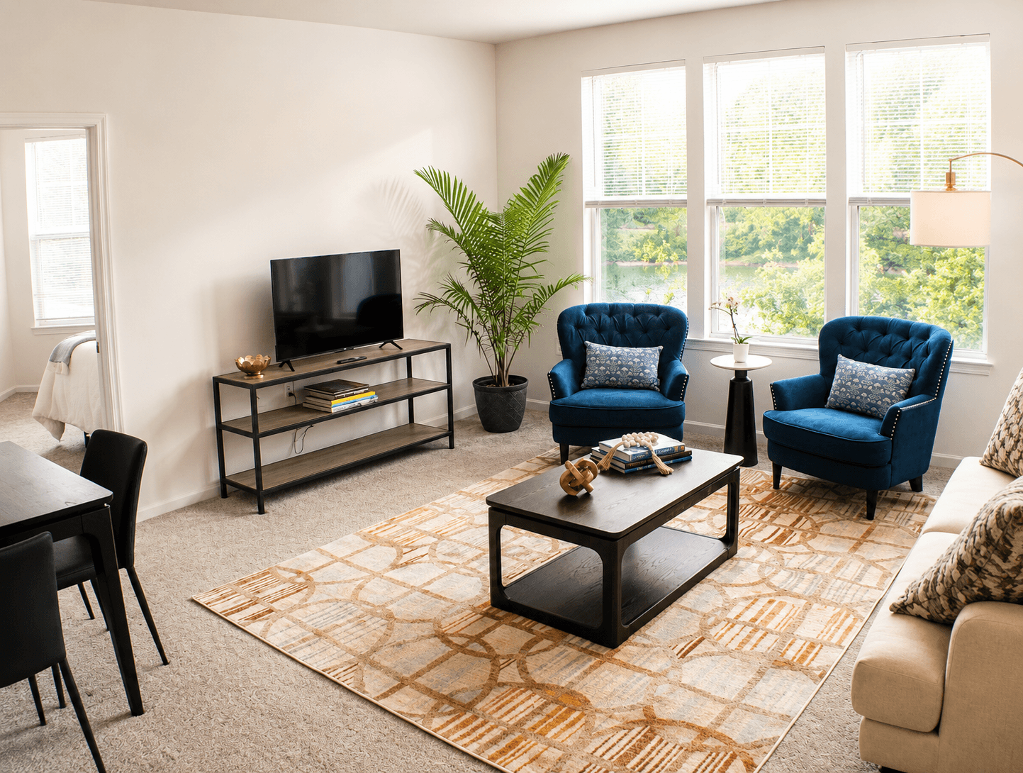 A cozy living room featuring two blue chairs, a coffee table, a TV stand, and large windows with a view of greenery.
