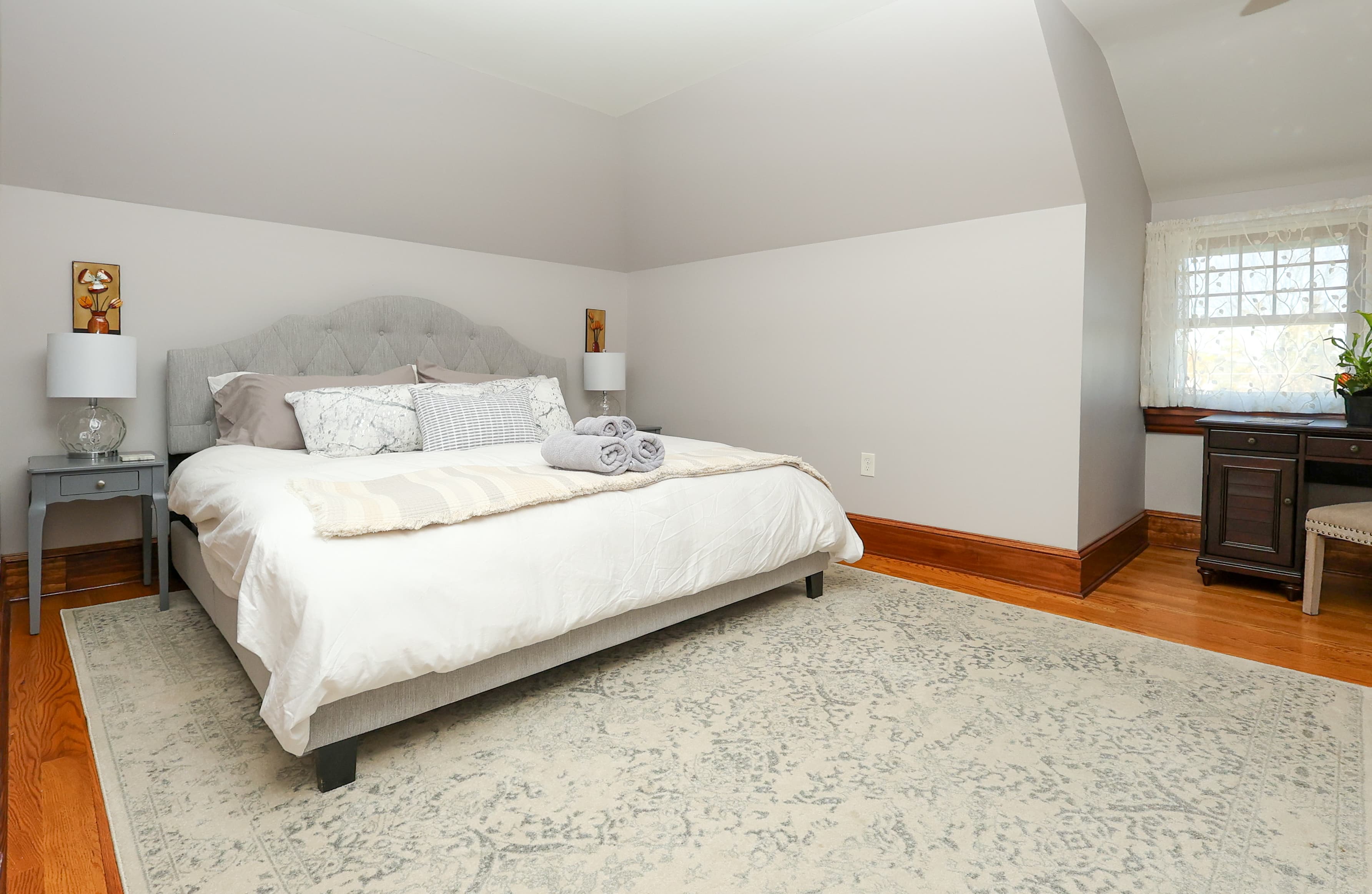 A cozy bedroom features a light gray upholstered bed, two bedside lamps, and a decorative rug on wooden flooring.