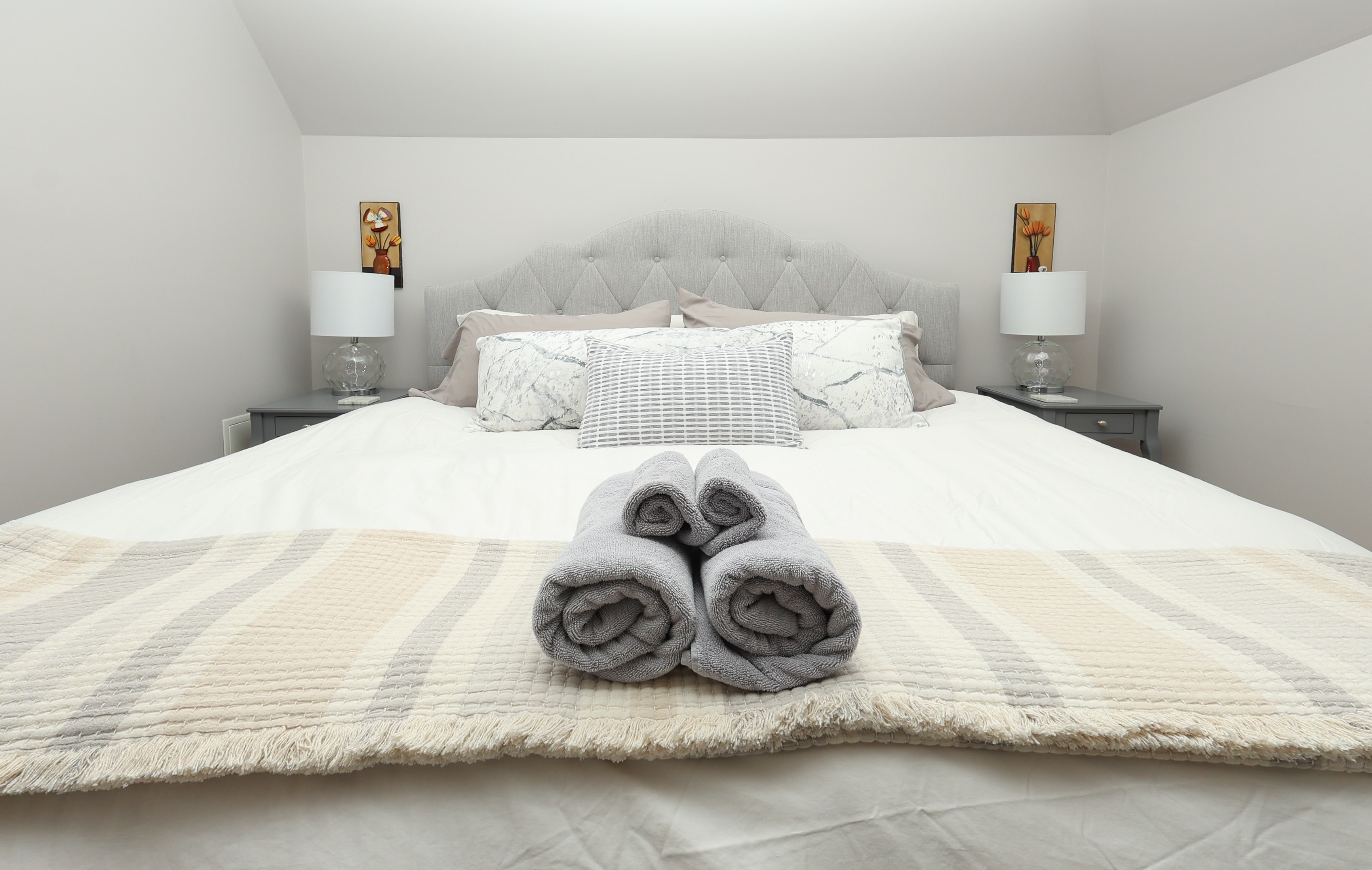A neatly made bed with gray towels stacked in the foreground, flanked by bedside lamps and decorative artwork.