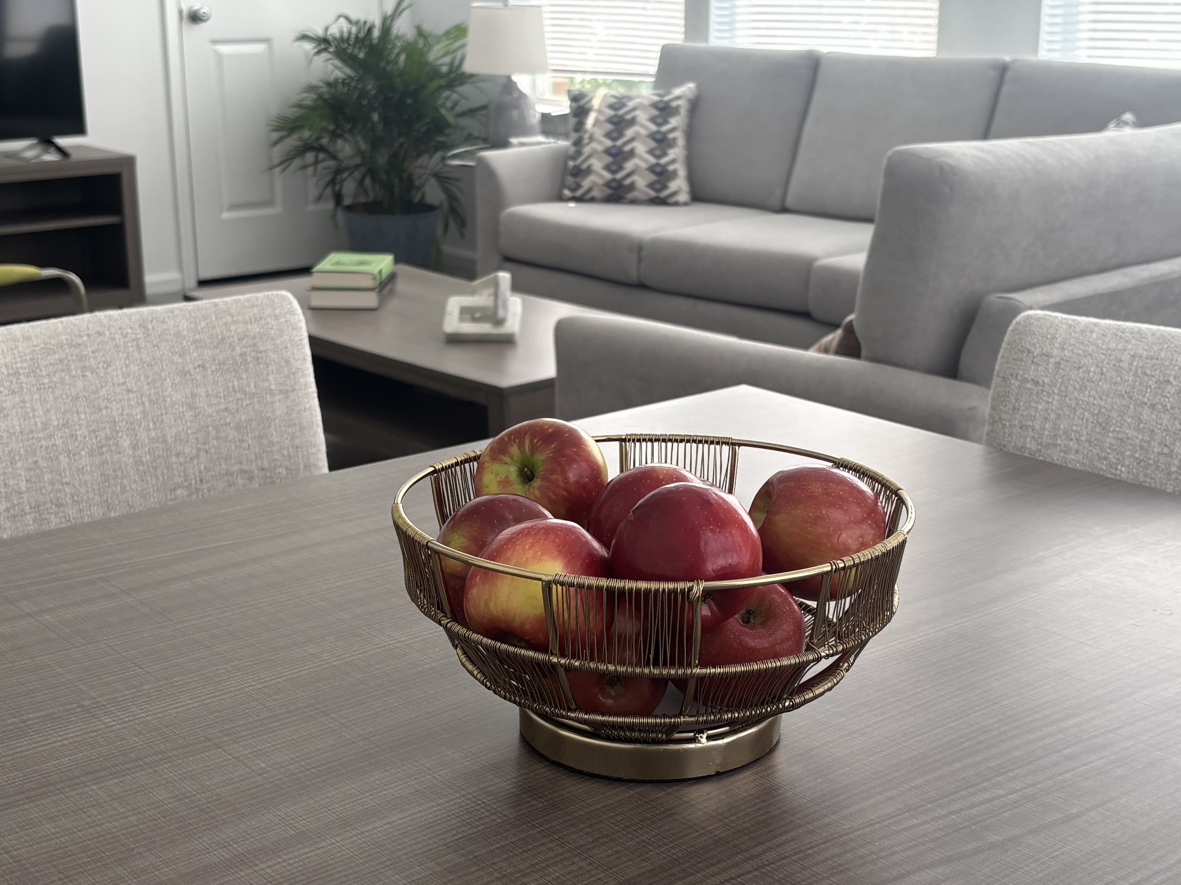 A bowl of red apples sits on a wooden dining table in a modern living space.