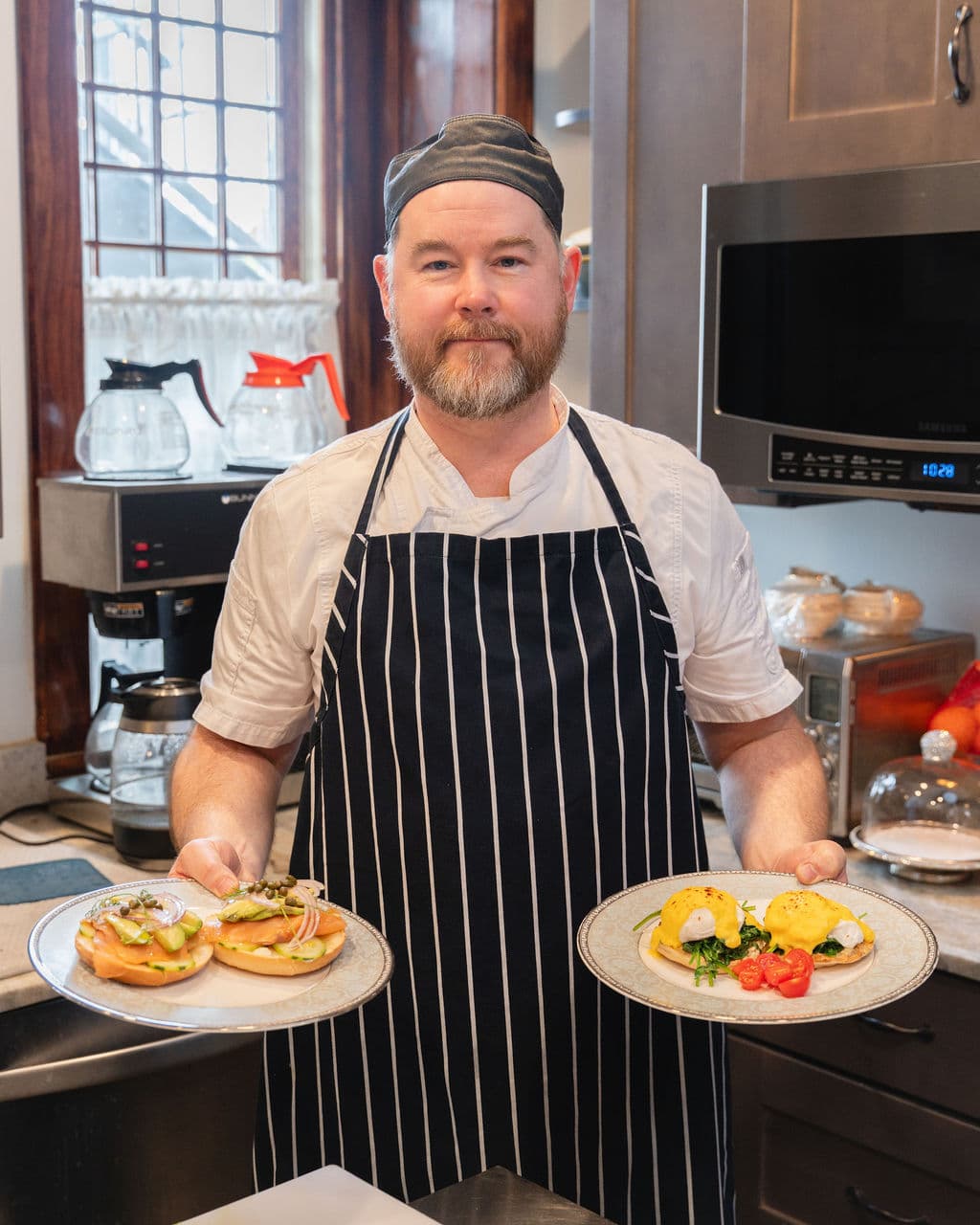 A chef in a striped apron holds two plates of gourmet food in a kitchen setting.