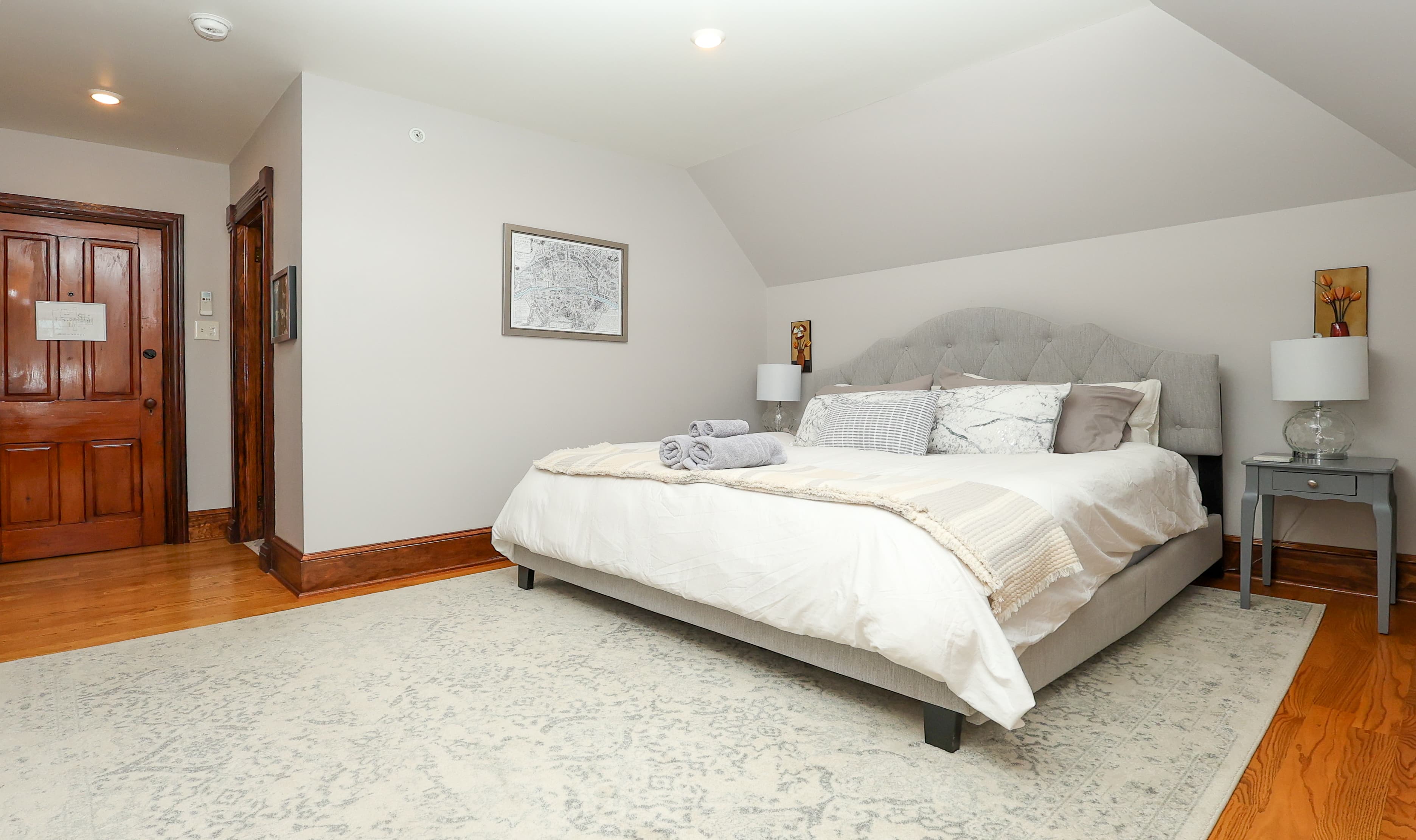 A cozy bedroom featuring a grey bed, two bedside lamps, a small table, and wooden doors.